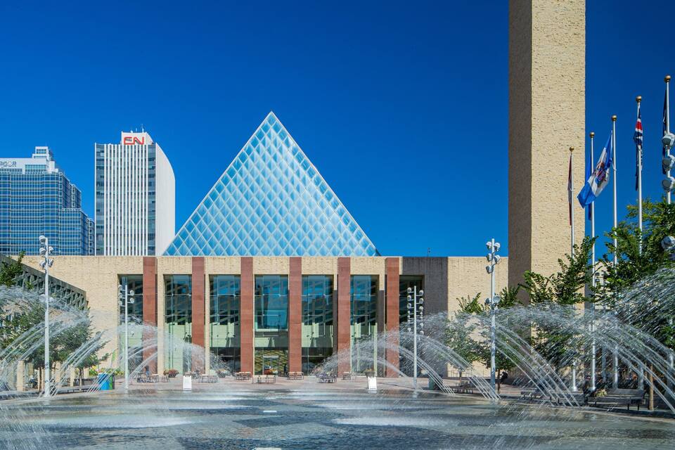 A modern building with a glass pyramid, surrounded by a water fountain and flags.