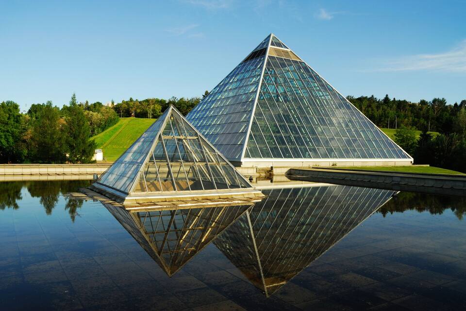 Two glass pyramids reflected in a body of water, surrounded by greenery and a clear sky.