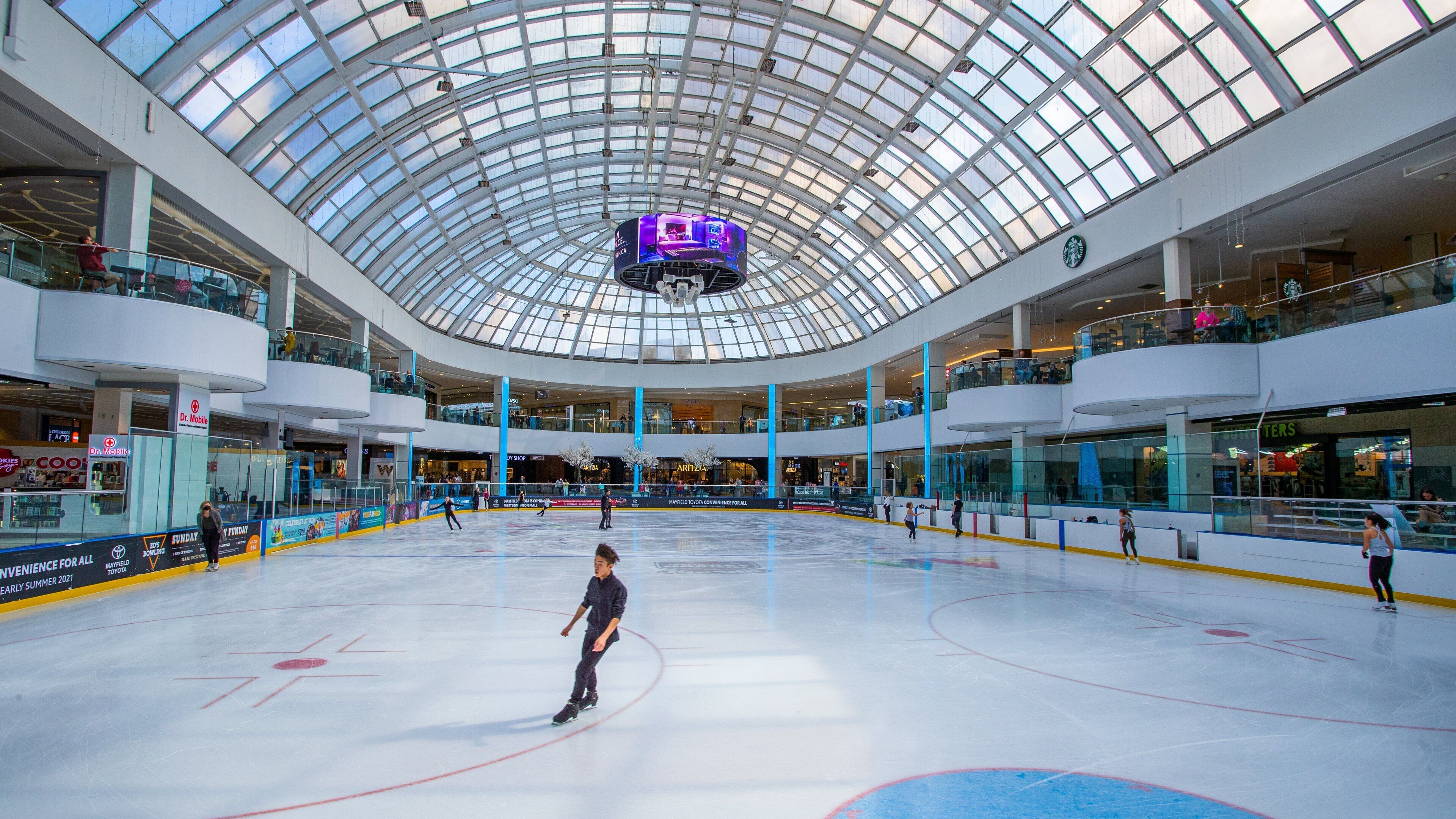 An indoor ice rink with a glass ceiling, surrounded by shopping malls and stores.