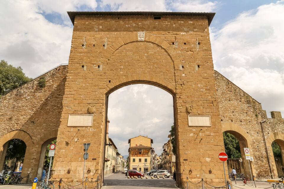 Un arc de pedra ornamentat amb un escut, flanquejat per edificis i un carrer amb cotxes i bicicletes aparcades.
