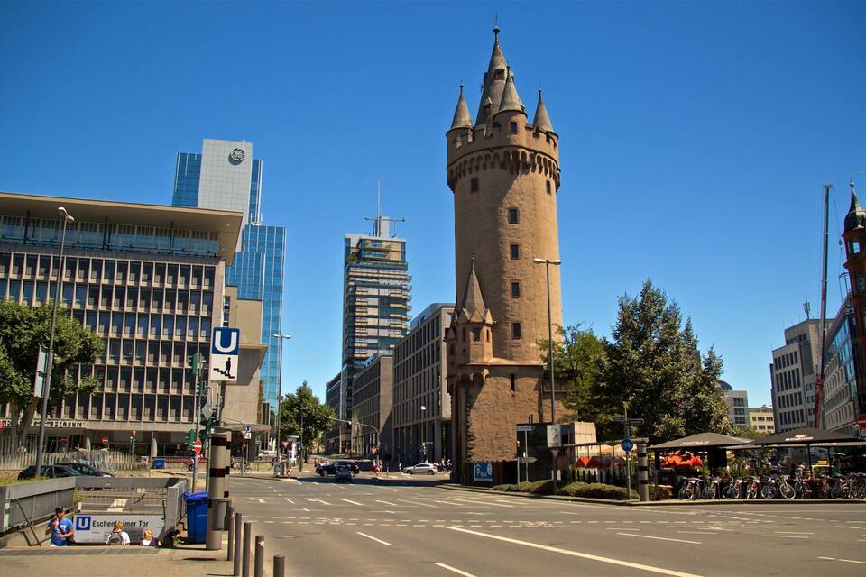 A city street with a historic tower, modern buildings, and a clear blue sky.