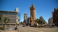 A city street with a historic tower, modern buildings, and a clear blue sky.
