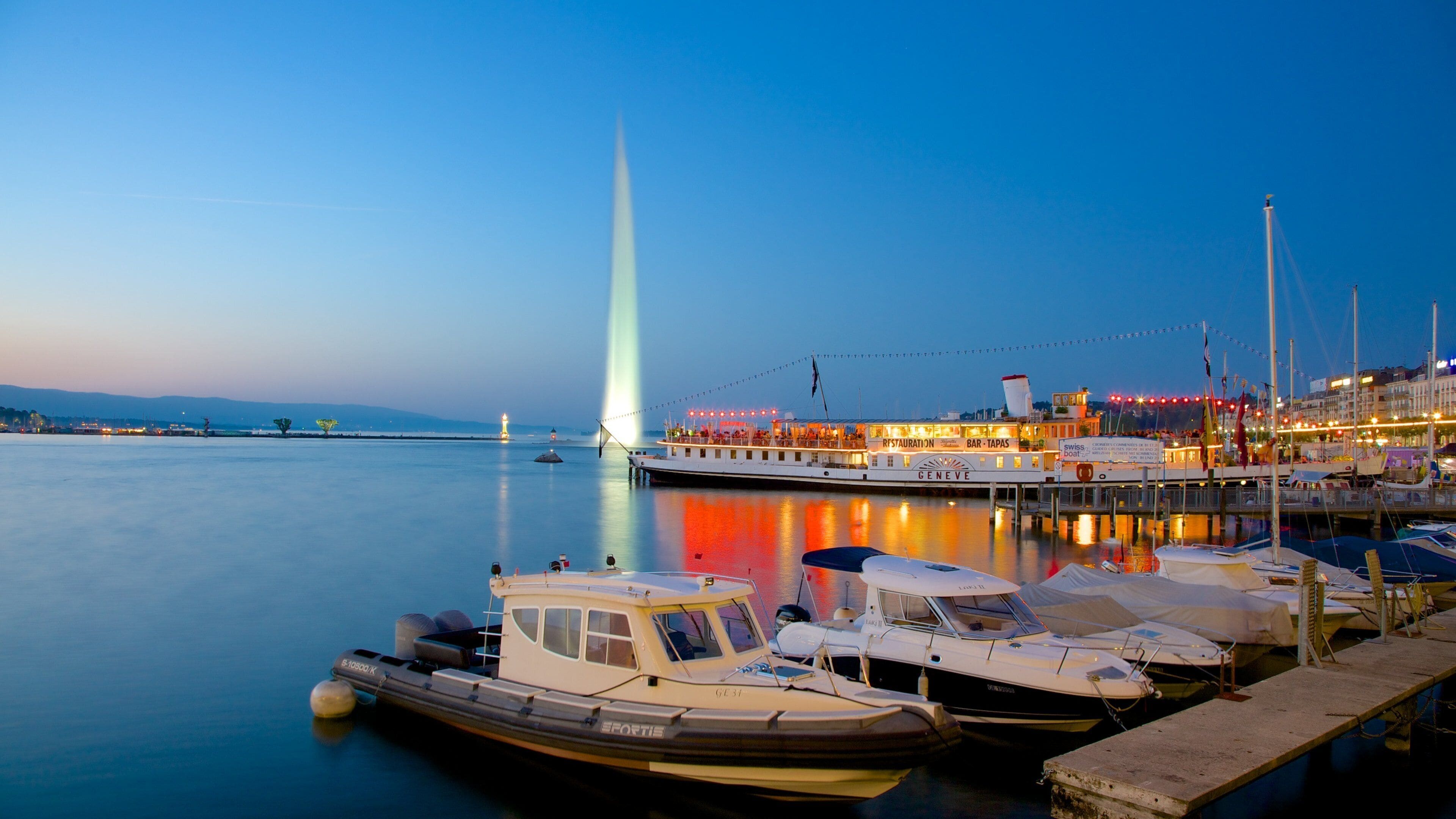 A marina at dusk with boats docked, a prominent light tower, and a restaurant named 'GENEVE' in the background.