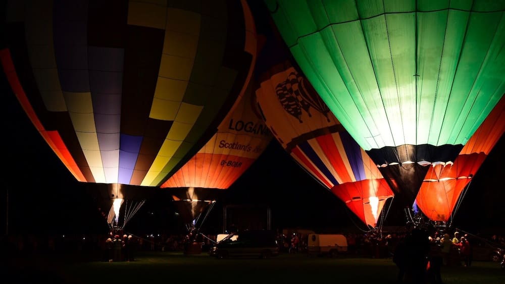 A nighttime hot air balloon festival with multiple balloons, some with visible logos and text.