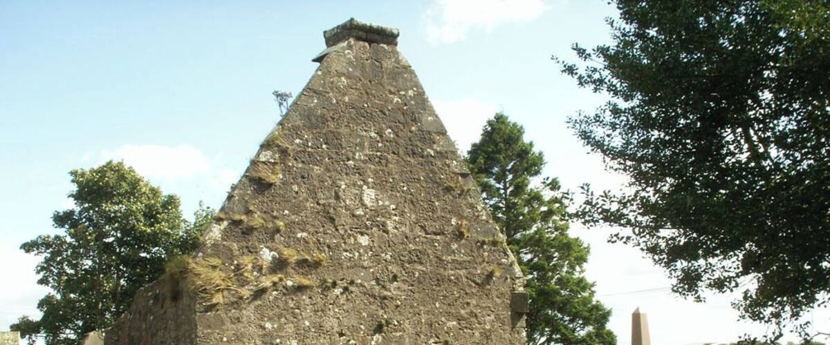 A historical stone structure with a pointed roof, surrounded by a cemetery with headstones.