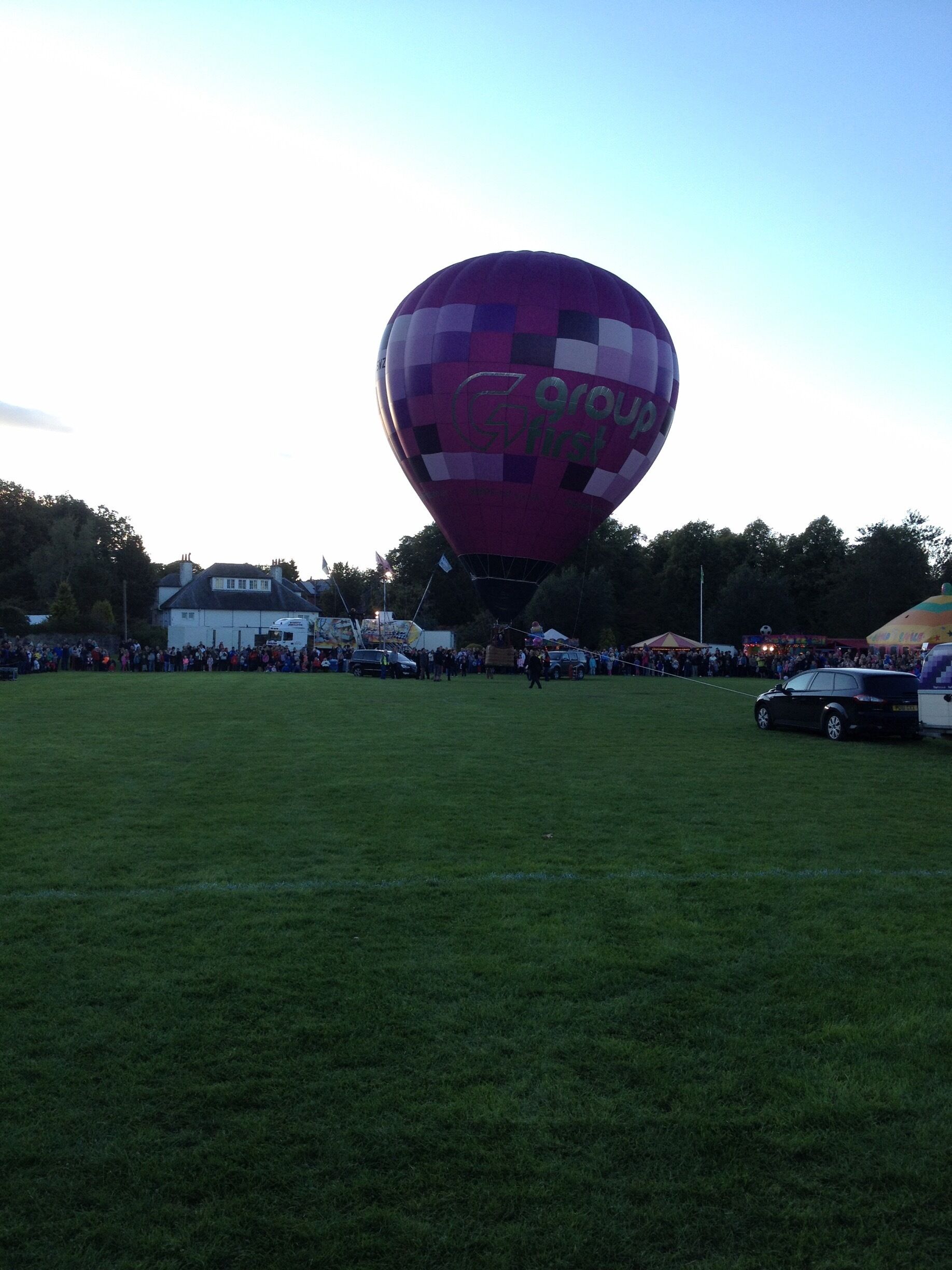 A large hot air balloon with the text "Groupp" on it, parked on a grassy field with a crowd of people and a car nearby.