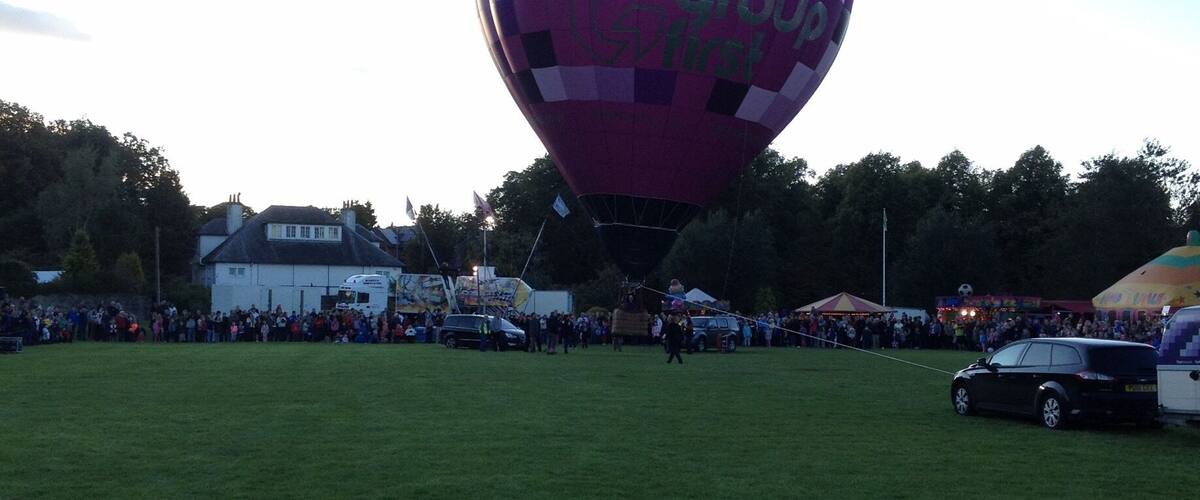 A large hot air balloon with the text "Groupp" on it, parked on a grassy field with a crowd of people and a car nearby.