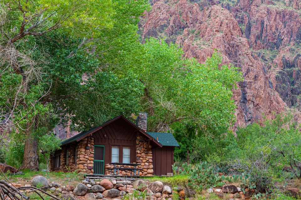 A rustic stone cabin surrounded by lush greenery and rocky cliffs.