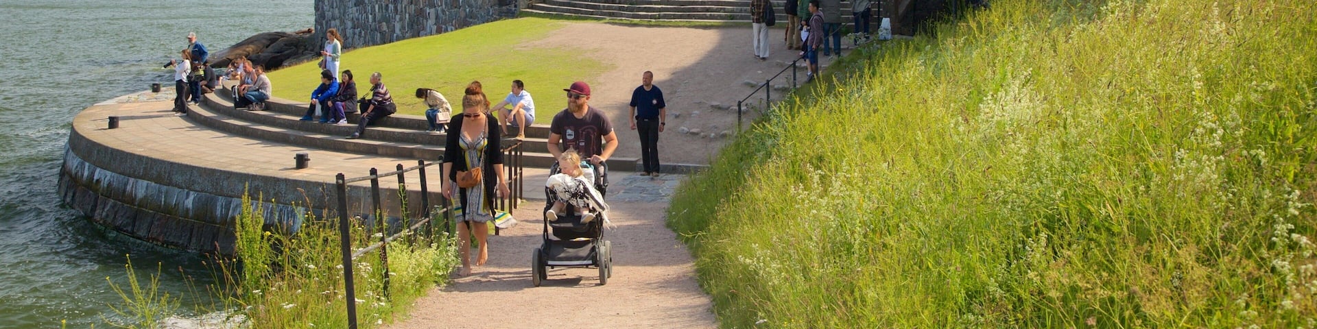 A stone fort with a grassy top, a circular viewing platform by the water, and a staircase leading up to the fort.