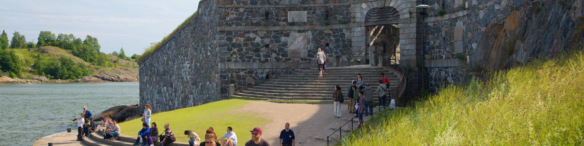 Uma fortaleza de pedra com um topo gramado, uma plataforma circular de observação perto da água e uma escadaria que leva à fortaleza.