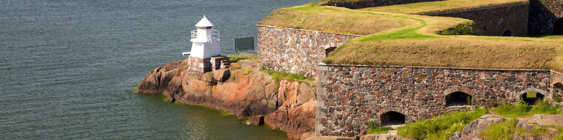 A white lighthouse on a rocky outcrop by the sea, with a stone fortification and grassy mounds.