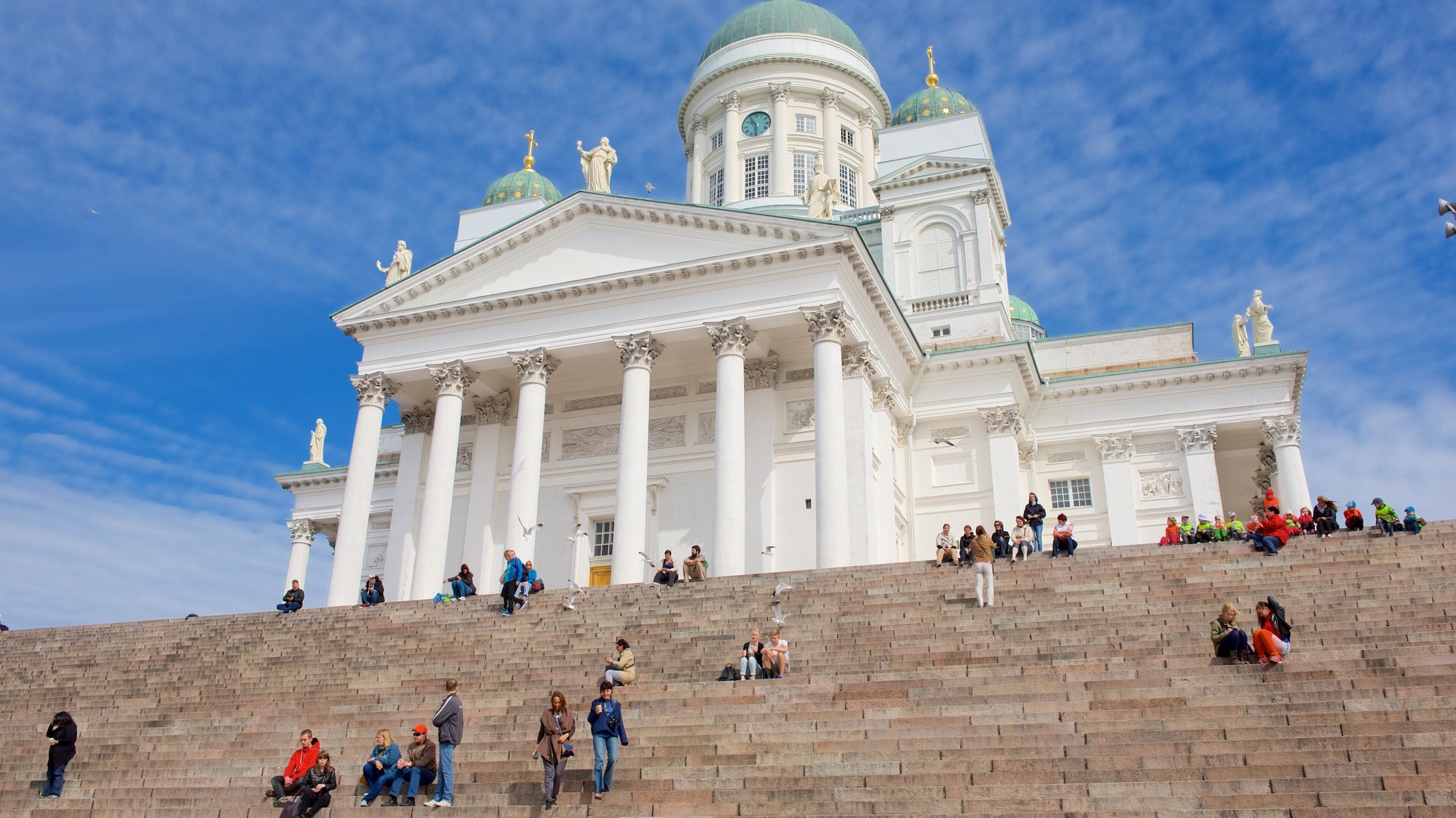 A grand white building with columns and a green dome, surrounded by people on the steps.