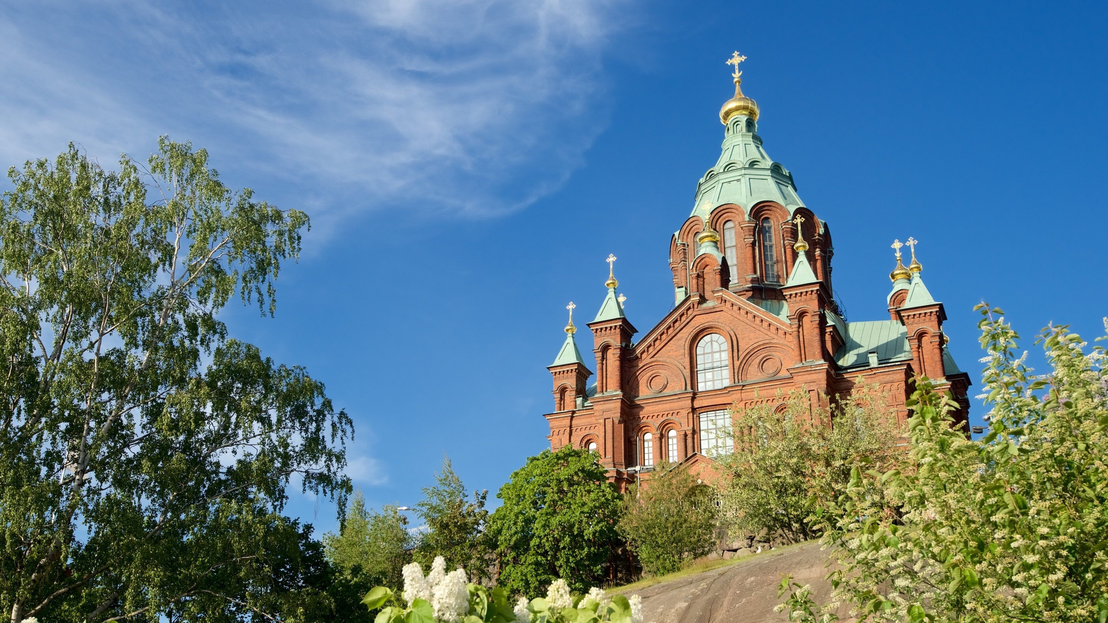 A large red brick church with green domes and crosses, surrounded by trees and flowers.