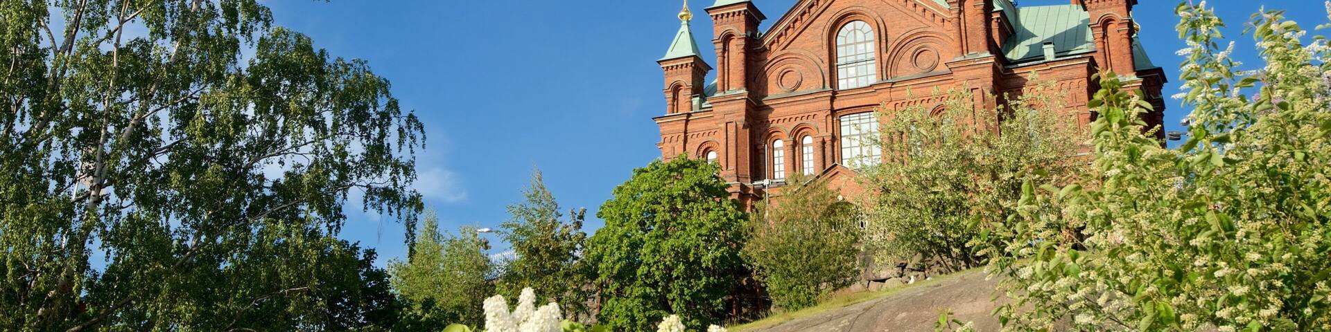 A large red brick church with green domes and crosses, surrounded by trees and flowers.