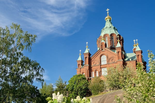 Eine große Kirche aus roten Backsteinen mit grünen Kuppeln und Kreuzen, umgeben von Bäumen und Blumen.