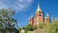 A large red brick church with green domes and crosses, surrounded by trees and flowers.