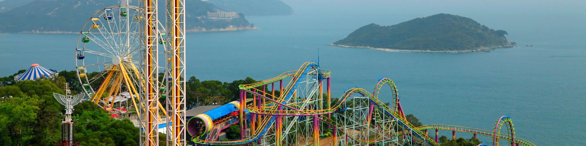 Um parque de diversões à beira-mar com roda gigante, montanhas-russas e uma vista panorâmica do mar e ilhas distantes.