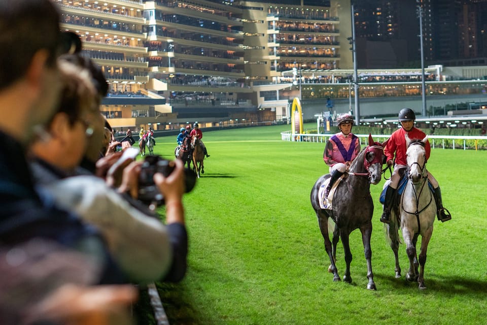 A horse race in progress at night with jockeys on horses and spectators in the stands.