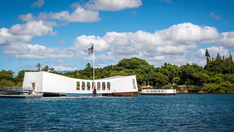 A white building with a flag on top, surrounded by water and greenery.