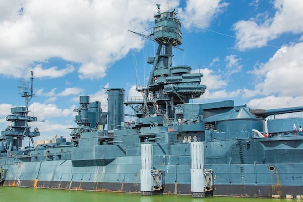 A large naval ship docked at a port with a clear sky and greenish water.