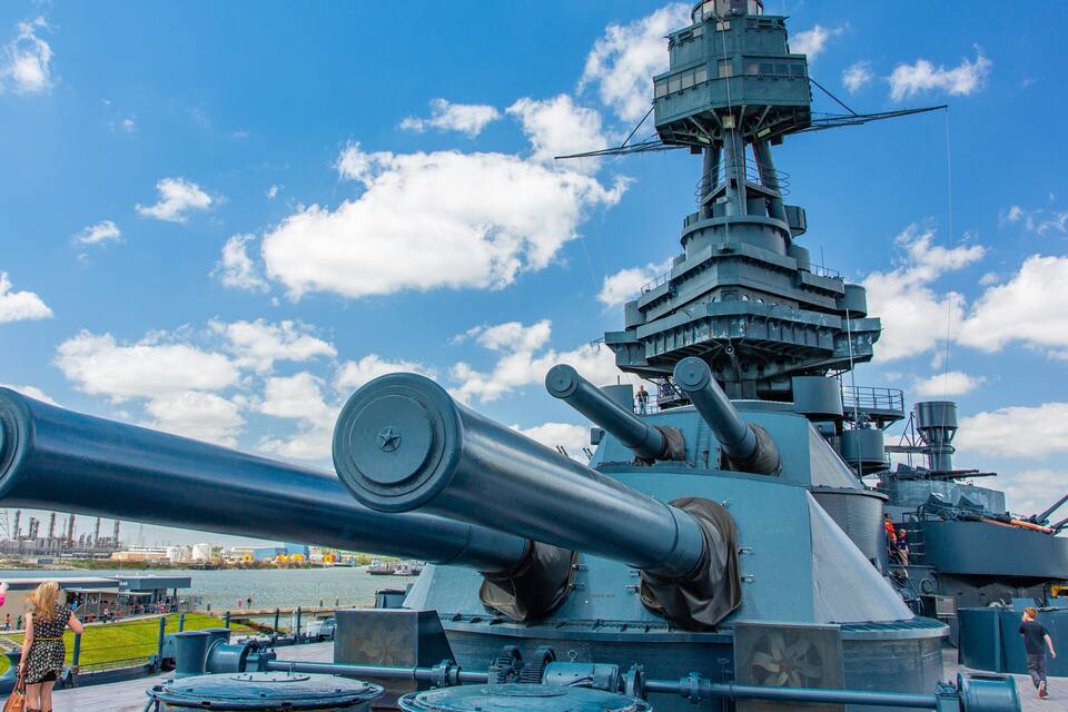 A large naval gun turret on a ship with a clear sky and clouds in the background.