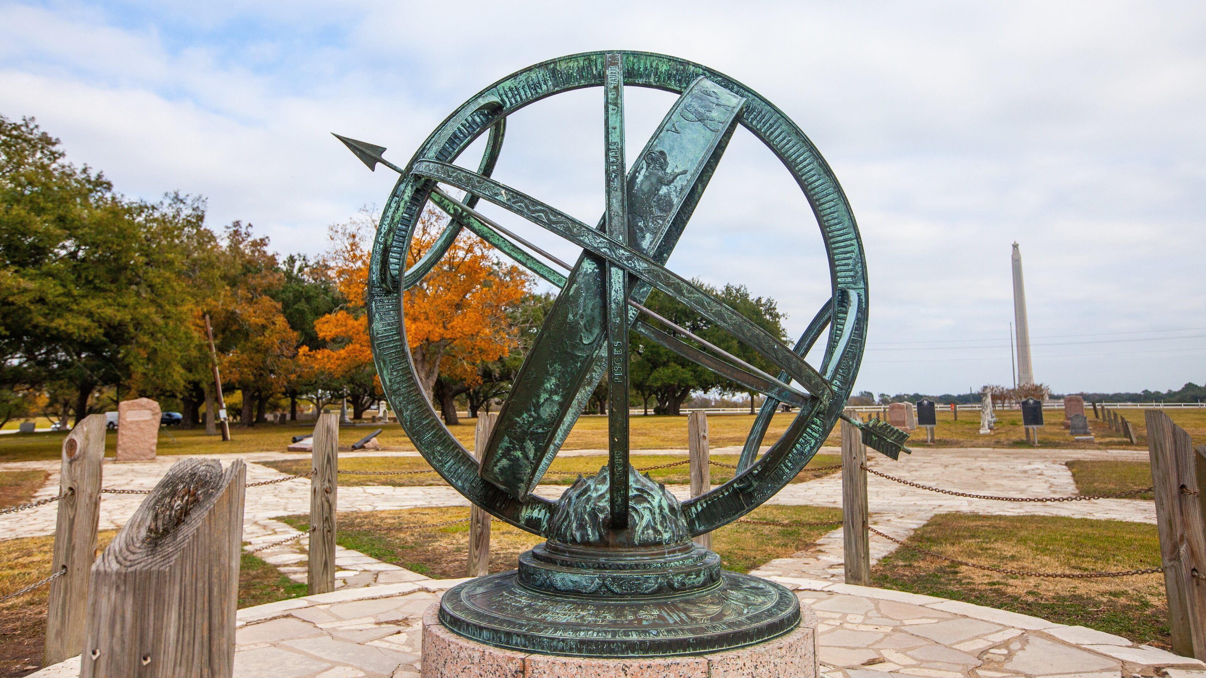 Una gran escultura de bronce de un astrolabio en un parque con árboles otoñales y un monumento distante.