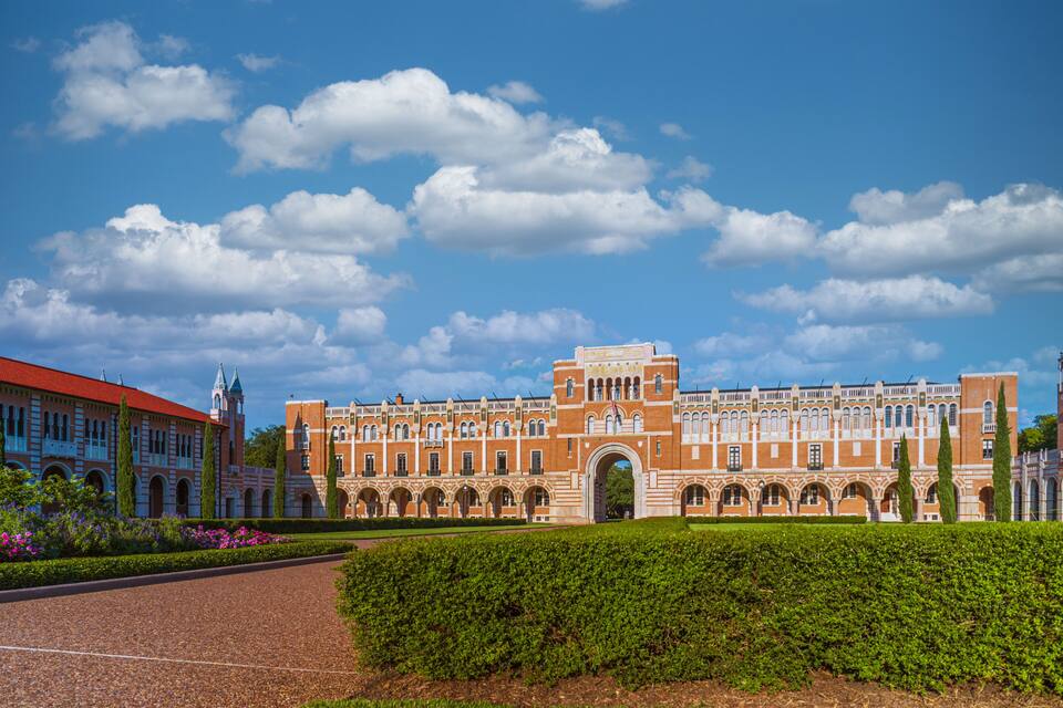 A large historic building with a red roof and arched windows, surrounded by a well-maintained garden and a clear blue sky.