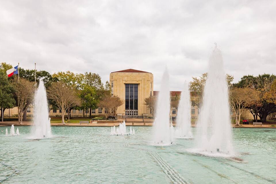 A fountain with multiple jets of water in front of a building with a flag.