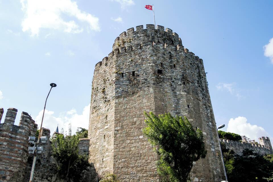 Una torre de pedra històrica amb una bandera, envoltada d'arbres i altres edificis antics.