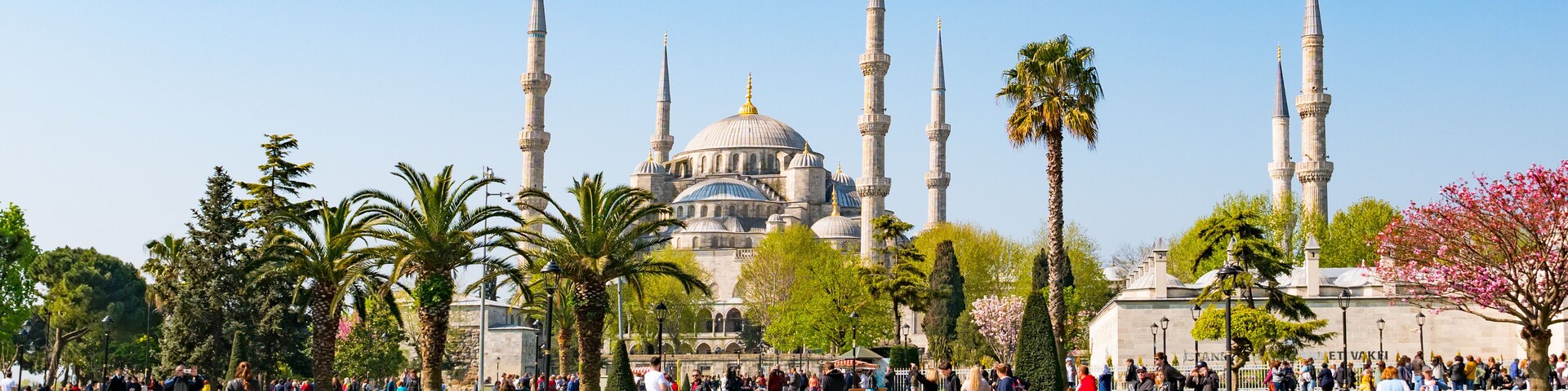 Uma mesquita histórica com múltiplos minaretes, uma cúpula central imponente e uma área de fontes ao redor, com pessoas.