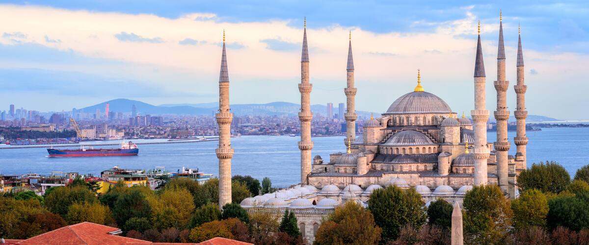 Une mosquée historique dotée de plusieurs minarets et de coupoles, une rivière et une vue sur la ville en arrière-plan.