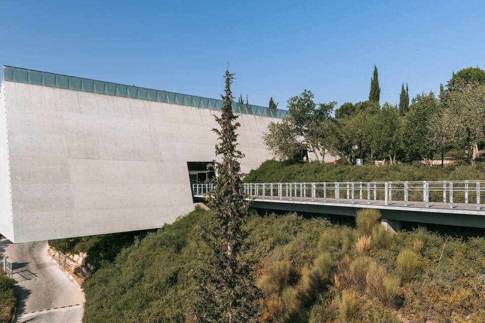 A modern concrete structure with a green roof, a pedestrian bridge, and surrounding greenery.