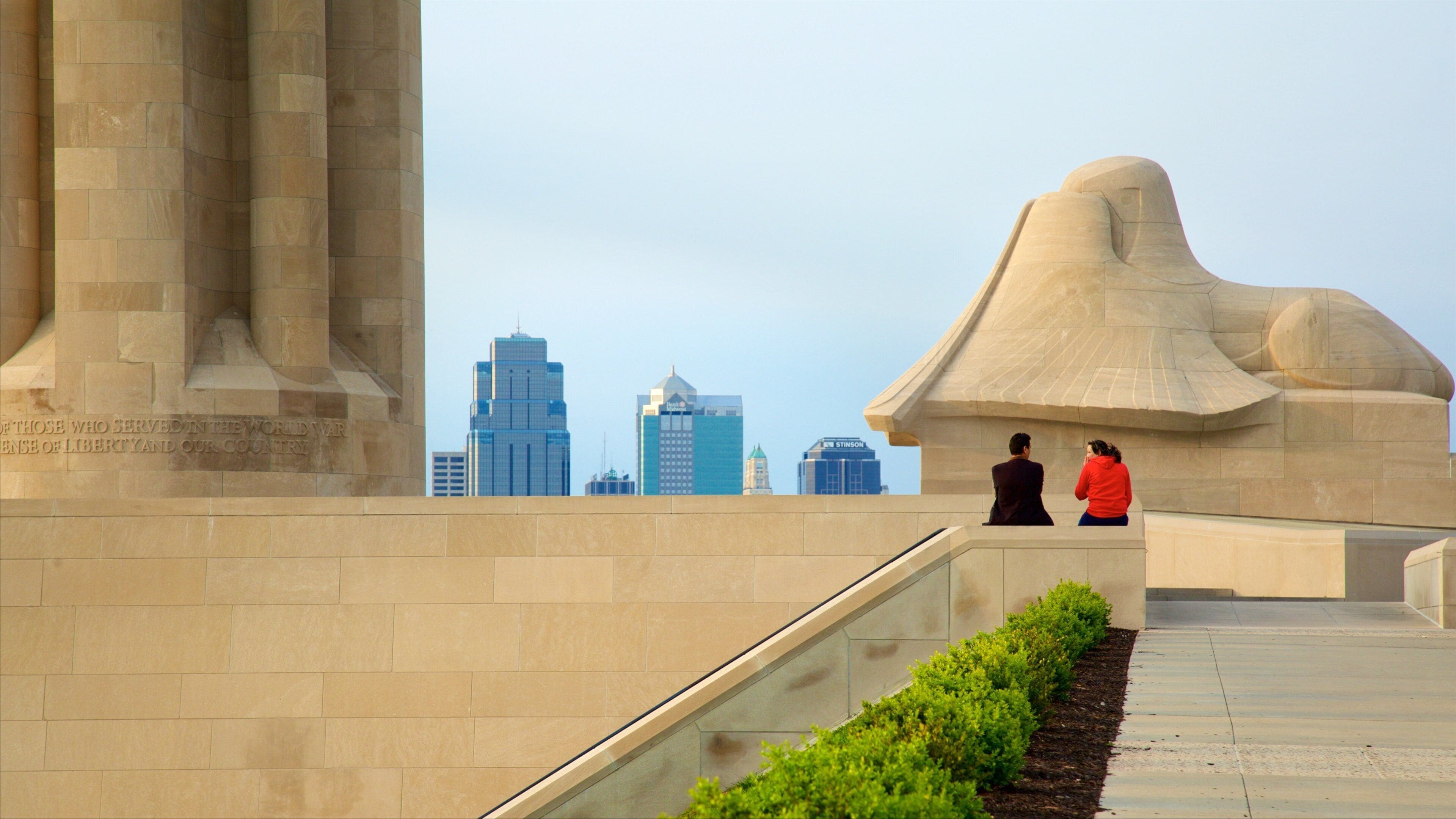 A monument with inscriptions, a city skyline, and a landscaped area with bushes.