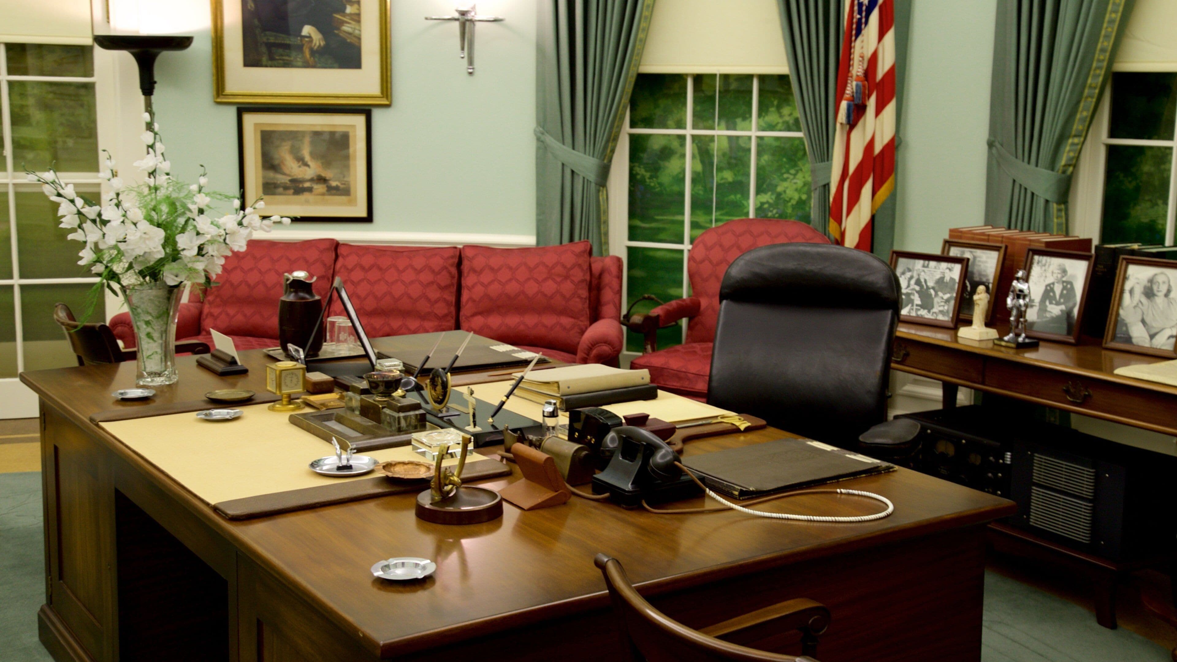 A formal office with a wooden desk, red upholstered chairs, framed pictures, and a vase with white flowers.