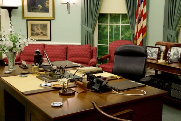 A formal office with a wooden desk, red upholstered chairs, framed pictures, and a vase with white flowers.