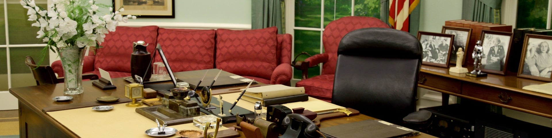 Escritório formal com mesa de madeira, cadeiras estofadas em vermelho, quadros emoldurados e um vaso com flores brancas.