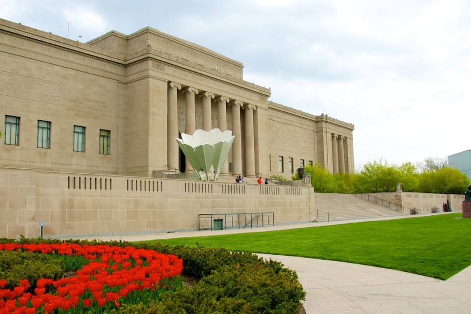 A large building with classical architecture, a central sculpture, and a well-maintained garden with red flowers.
