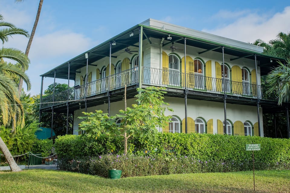A two-story colonial-style building with a balcony, surrounded by lush greenery and palm trees.