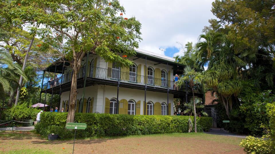 A historic building with green shutters and a balcony, surrounded by lush greenery and palm trees.