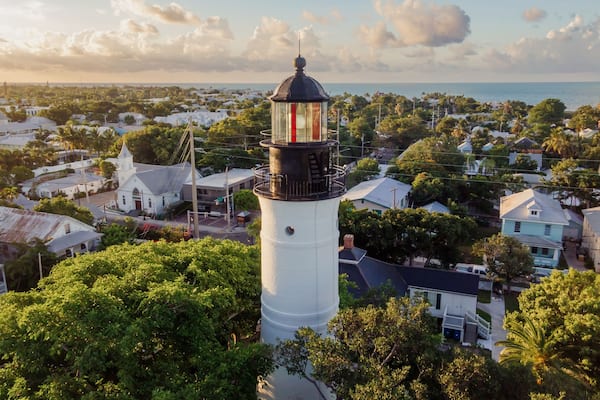 A white lighthouse with a black top, surrounded by lush greenery and residential buildings.