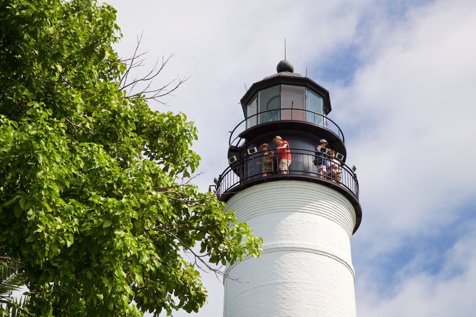 A lighthouse with a white exterior and a black top, surrounded by lush green trees.