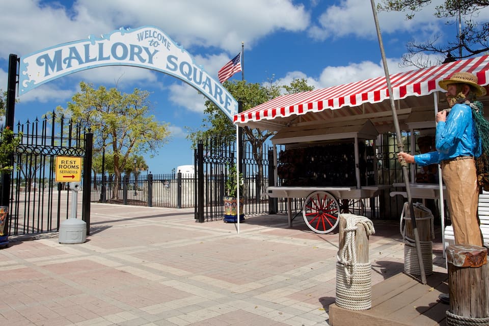 Cartell amb la frase 'Welcome to Mallory Square', una bandera dels Estats Units, un told retractable vermell i blanc, i una estàtua d'un home amb camisa blava i barret de palla.