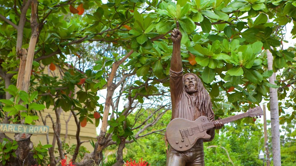 A statue of a musician with a raised arm and a guitar, set against a backdrop of green foliage and a building with a sign reading "MARLEY ROAD".