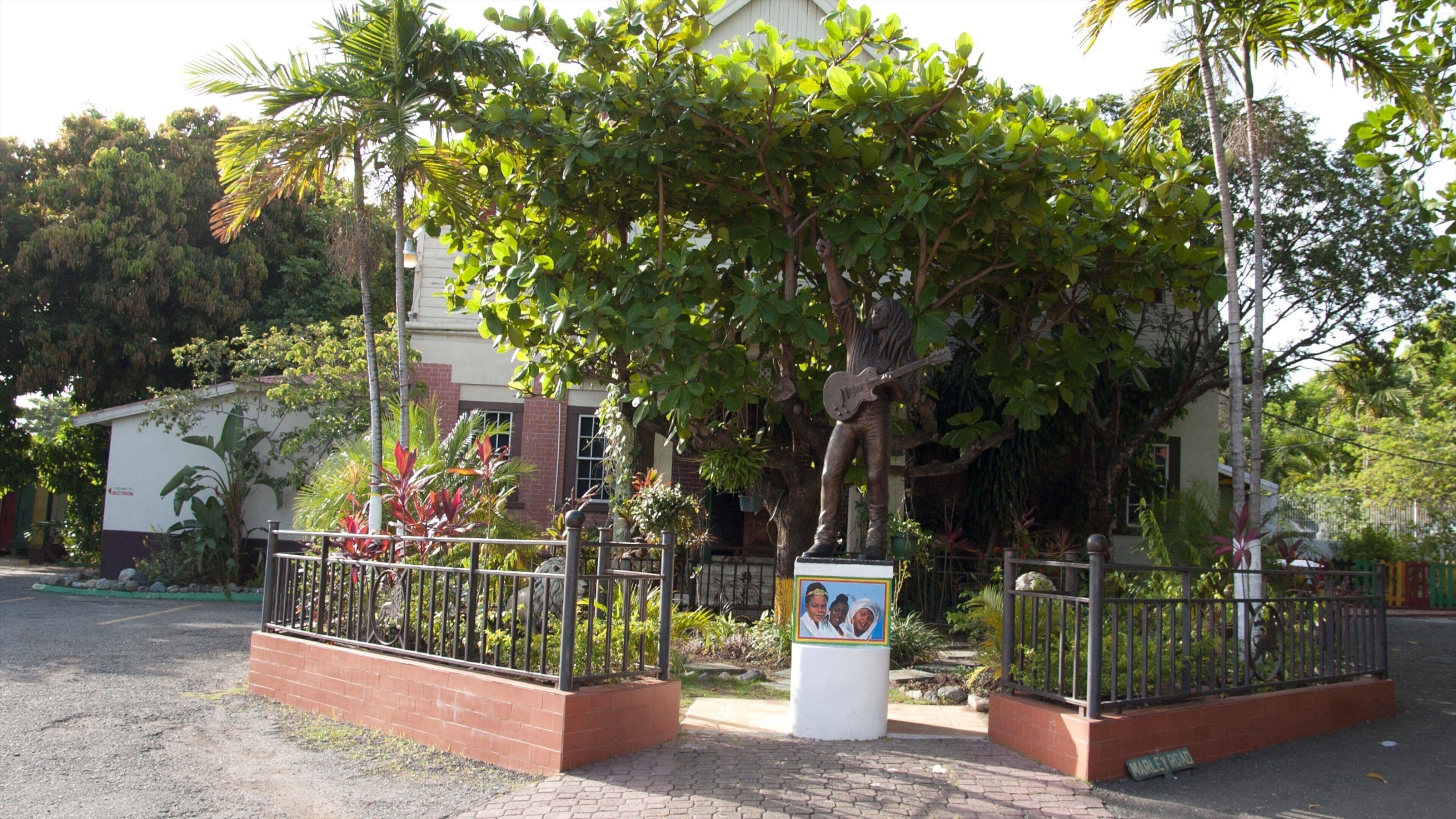 Jardim cercado com uma estátua de um homem tocando violão, rodeado por vegetação exuberante e um edifício ao fundo.