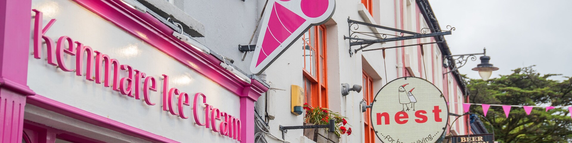 Fachada rosa com o nome Kenmare Ice Cream, um letreiro com a palavra 'nest' e uma ilustração de pássaro, e um letreiro de 'Beer Garden'.
