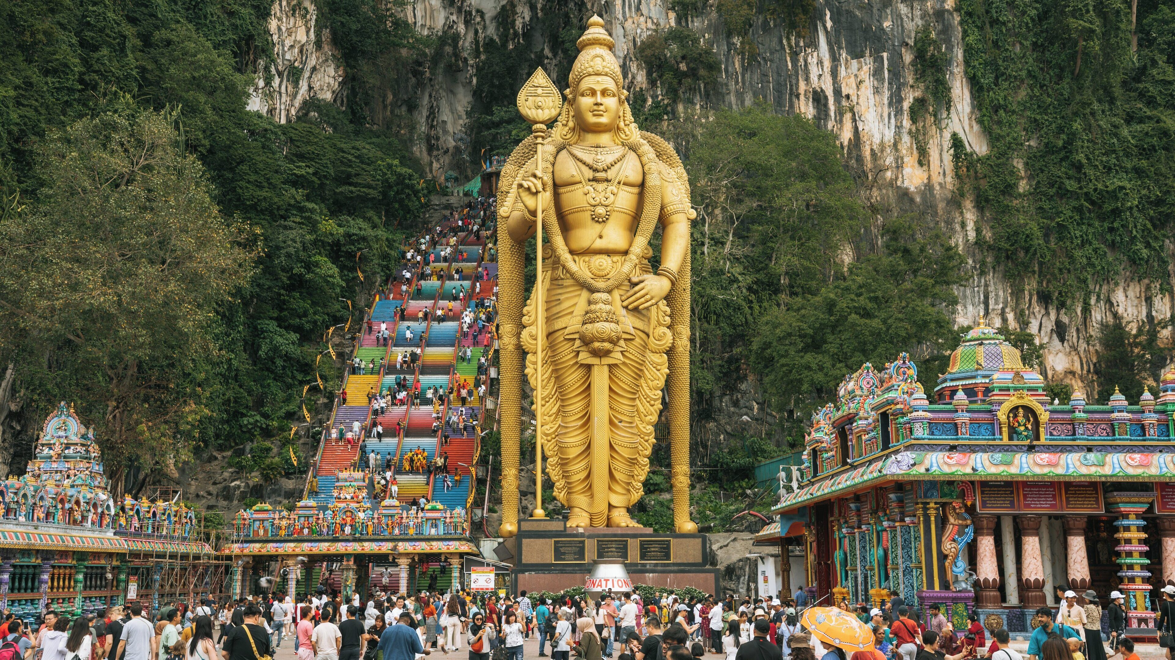 Golden statue stands majestically at Batu Caves in Kuala Lumpur, surrounded by colorful steps and vibrant temple architecture during a bustling visit