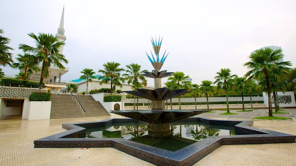 A fountain with a modern design, surrounded by palm trees and a mosque with a tall minaret in the background.
