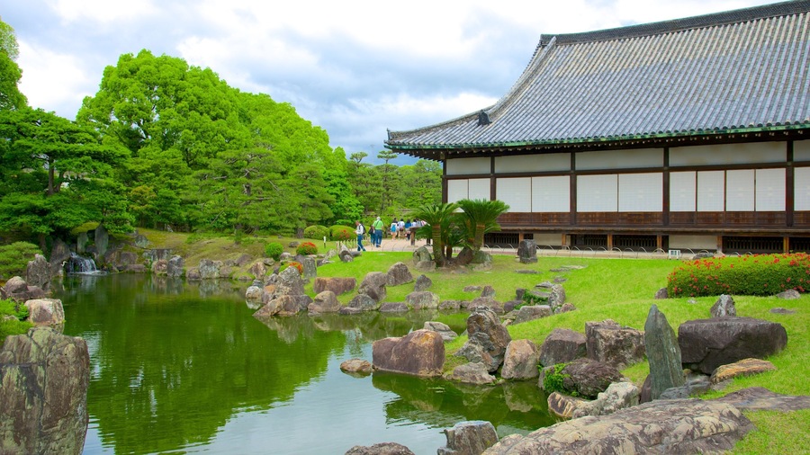 A traditional Japanese building with a tiled roof, surrounded by lush greenery and a calm pond with rocks.