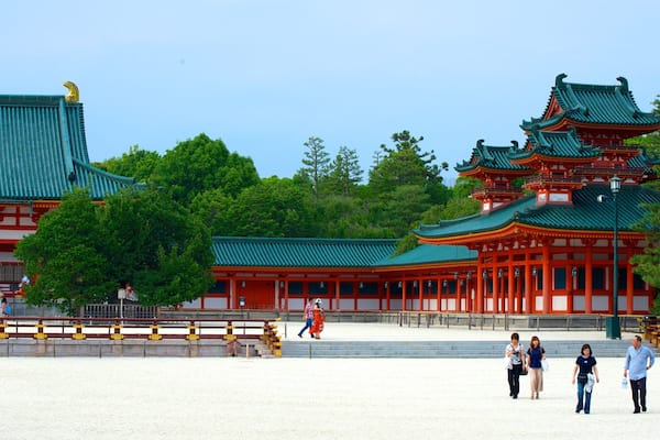 Um templo japonês tradicional com telhados vermelhos e verdes, rodeado por árvores e uma área arenosa com pessoas a passear.