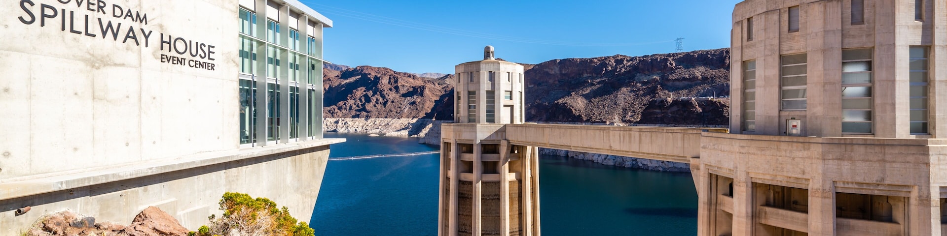 Uma barragem com uma descarga identificada como 'HOOVER DAM SPILLWAY HOUSE EVENT CENTER' e um céu azul límpido.